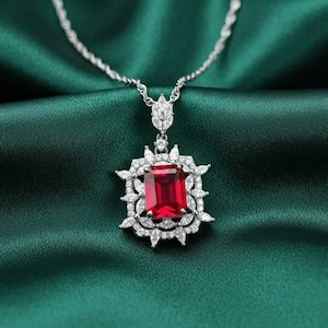 May include: A silver necklace with a large, rectangular, red gemstone pendant. The pendant is surrounded by a halo of smaller, clear gemstones and leaf-shaped accents. The necklace rests on a dark green satin fabric.