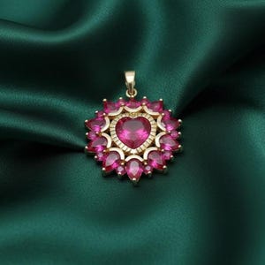 May include: A gold-toned pendant with a heart-shaped, ruby-colored center stone, surrounded by smaller, teardrop-shaped stones. The pendant is set against a dark green, silky background, creating a luxurious feel.