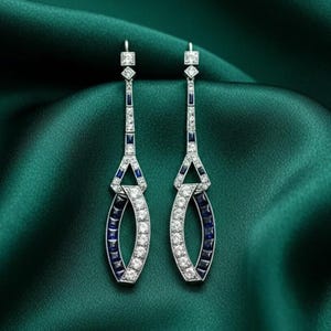 May include: A pair of Art Deco-style earrings with a geometric design. The earrings feature a combination of white and blue gemstones set in a silver-toned metal. The elongated shape and intricate detailing create a vintage aesthetic.