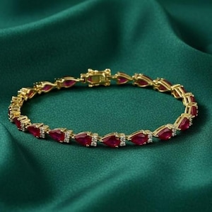 May include: A gold-toned bracelet with a series of teardrop-shaped, deep red gemstones. Small, clear stones are interspersed between the red gems. The bracelet is set against a dark green, textured background.
