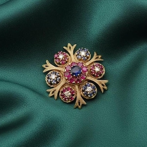 May include: A gold brooch with a floral design. The brooch features a central blue gemstone surrounded by red gemstones and smaller clusters of red, blue, and white gemstones. The brooch is set against a dark green fabric.