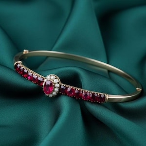 May include: Gold-toned bangle bracelet with a central oval ruby surrounded by small pearls. The bracelet features a row of red gemstones and is set against a dark green satin background. A classic piece of jewelry.