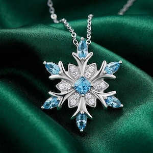 Könnte beinhalten: Eine silberne Schneeflocken-Anhänger-Halskette mit blauen und weißen Edelsteinen. Der Anhänger hat einen zentralen quadratischen blauen Stein, umgeben von kleineren weißen Steinen und sechs blauen tropfenförmigen Steinen. Die Halskette liegt auf grünem Satin.