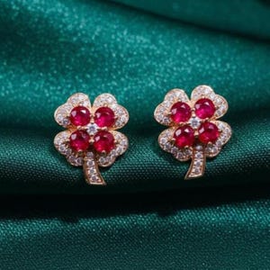 May include: A pair of gold-toned clover-shaped stud earrings. Each earring features four vibrant red gemstones surrounded by small, clear crystals. The earrings are set against a dark green, textured background.