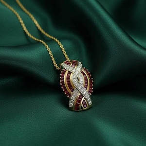 May include: A gold necklace with a pendant featuring a design of red gemstones, diamonds, and gold accents. The pendant is suspended from a delicate gold chain. The necklace is displayed on a dark green satin fabric.