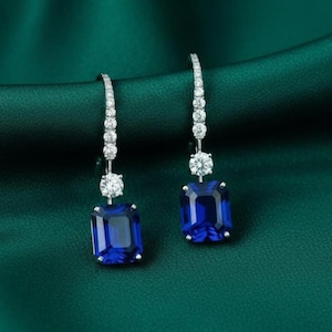 May include: A pair of elegant drop earrings featuring a vibrant blue emerald-cut gemstone. The earrings have a delicate design with a row of small, sparkling white stones leading to a larger round white stone, and a large blue gemstone. The earrings are set against a dark green background.