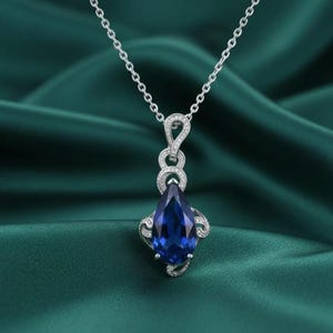 Könnte beinhalten: Eine silberne Halskette mit einem tropfenförmigen Anhänger aus tiefblauem Edelstein. Der Anhänger ist mit kleinen, klaren Steinen verziert und hängt an einer filigranen Kette. Der Hintergrund ist ein dunkelgrüner, strukturierter Stoff.
