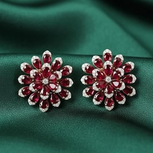 Può includere: Un paio di orecchini a forma di fiore. Ogni orecchino presenta un gruppo di pietre preziose rosso scuro circondate da pietre preziose bianche più piccole. Gli orecchini sono appoggiati su uno sfondo verde scuro e serico.