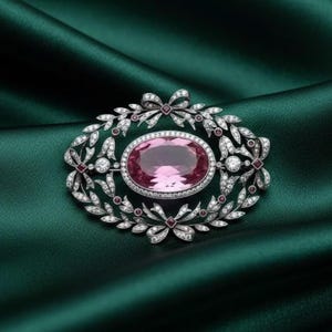 May include: An oval-shaped brooch with a large pink gemstone at its center, surrounded by a silver-toned frame. The frame features a leaf and bow design, accented with small diamonds and red gemstones. The brooch is set against a dark green background.
