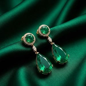 Pode incluir: Um par de brincos de ouro com grandes pedras preciosas verde esmeralda. Cada brinco apresenta uma esmeralda redonda rodeada por pequenos diamantes no topo, com uma esmeralda em forma de gota pendurada por baixo. Fundo de cetim verde escuro.