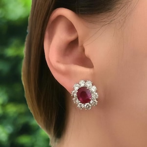 May include: A close-up of a stud earring. It features a large, oval, deep red gemstone, encircled by smaller, clear gemstones. The earring is set in silver-coloured metal and is worn on an ear.