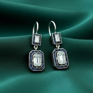 Könnte beinhalten: Ein Paar Ohrhänger im Art-Deco-Stil. Jeder Ohrring hat einen rechteckigen, klaren Edelstein, der in einen dunkelblauen Rahmen gefasst ist, mit einem kleineren passenden Stein darüber. Die Ohrringe hängen an einem silbernen Haken.