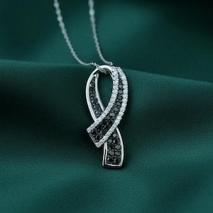 May include: A silver pendant necklace featuring a ribbon design. The ribbon is adorned with black and white gemstones, creating a striking contrast. The necklace hangs on a delicate silver chain, set against a dark green background.