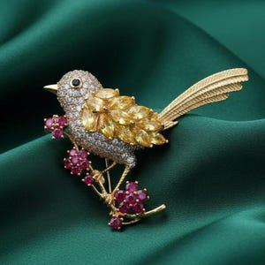 May include: A gold-toned bird brooch with a detailed design. The bird's body is encrusted with clear and yellow gemstones, with ruby-colored flowers on a branch. The brooch has a textured gold tail and is set against a dark green background.