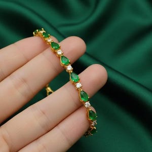May include: Gold bracelet featuring pear-shaped green gemstones and small, round, clear gemstones. The bracelet is set against a dark green background, highlighting the jewelry's sparkle and design. A delicate piece of jewelry.
