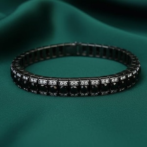 Puede incluir: Un brazalete negro con piedras negras cuadradas y pequeñas piedras blancas. El brazalete está sobre un fondo verde oscuro, creando un contraste llamativo. Es una pieza de joyería.