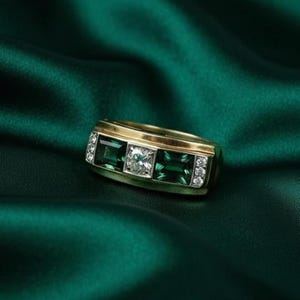 Könnte beinhalten: Ein goldener Ring mit rechteckigem Design, der drei smaragdgeschliffene grüne Edelsteine und einen zentralen quadratischen Diamanten aufweist. Kleine Diamanten sind auf beiden Seiten der grünen Steine gesetzt. Der Ring ist vor einem dunkelgrünen Stoff platziert.