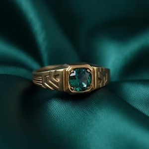 May include: A gold ring featuring a square, emerald-green gemstone. The ring's band has geometric patterns. The gemstone is set in a square bezel, and the ring is shown against a dark green, textured backdrop.