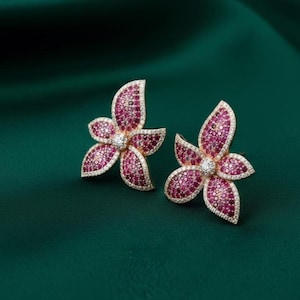 Pode incluir: Um par de brincos de botão em forma de flor. Cada brinco apresenta três pétalas, com uma pedra central transparente e uma borda de pedras transparentes menores. As pétalas são preenchidas com pequenas pedras rosa, fixadas em um metal dourado.