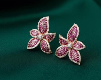May include: A pair of flower-shaped stud earrings. Each earring has three petals, with a central clear stone and a border of smaller clear stones. The petals are filled with small, pink stones, set against a gold-coloured metal.