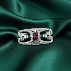 Puede incluir: Broche plateado de forma rectangular, adornado con numerosos diamantes pequeños y brillantes. El centro presenta un vibrante rubí de corte ovalado. El broche se encuentra sobre un fondo de tela de satén verde esmeralda.