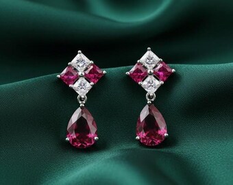 May include: A pair of drop earrings with a square cluster of pink and white gemstones above a teardrop-shaped, pink gemstone. The earrings are set against a dark green, silky background.