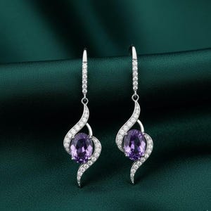 Op de afbeelding: Een paar zilverkleurige oorhangers. Elke oorbel heeft een grote, ovale, paarse edelsteen, omringd door een wervelend ontwerp van kleinere, heldere edelstenen. De oorbellen hangen aan een eenvoudige haak.