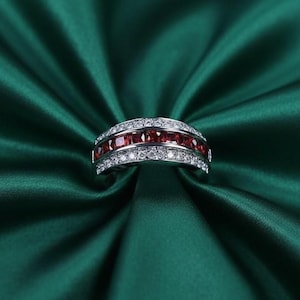 May include: A silver ring with a band of square-cut red gemstones, bordered by rows of smaller, clear gemstones. The ring is set against a backdrop of deep green fabric, creating a striking contrast.
