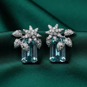 Könnte beinhalten: Ein Paar elegante Ohrringe mit rechteckigen, hellblauen Edelsteinen. Jeder Ohrring hat ein Blumendesign mit funkelnden, klaren Steinen und Blattakzenten, vor einem dunkelgrünen Hintergrund.