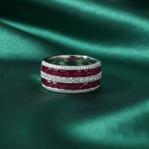 May include: A silver ring with three rows of gemstones. The top and bottom rows feature square-cut, deep red gemstones, while the middle row has round, clear gemstones. The ring is set against a dark green background.