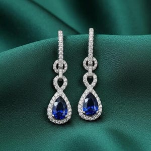 Puede incluir: Un par de elegantes pendientes colgantes. Cada pendiente presenta una piedra preciosa azul oscuro en forma de pera, rodeada por un halo de pequeñas piedras brillantes. Los pendientes tienen un delicado diseño en forma de infinito.