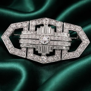 May include: A dazzling, Art Deco-style brooch. The silver-toned metal setting is encrusted with numerous sparkling diamonds. The design features geometric shapes and a central, round diamond. The brooch is set against a deep green satin background.