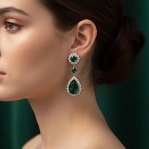May include: A pair of drop earrings with emerald green gemstones. The earrings have a floral design at the top, a round stone in the middle, and a pear-shaped stone at the bottom, all surrounded by a halo of small, clear stones. The earrings are set in a silver-coloured metal.