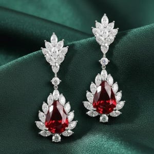 Puede incluir: Un par de elegantes pendientes colgantes con una gran gema roja en forma de lágrima, rodeada de cristales transparentes. Los pendientes tienen un engaste de metal plateado y un diseño en forma de hoja.