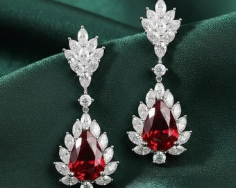 May include: A pair of drop earrings with a large, teardrop-shaped, deep red gemstone surrounded by clear crystals. The earrings have a silver-coloured metal setting and a leaf-like design. Ideal for formal events.