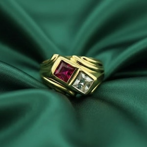 May include: A gold ring with two square gemstones, one red and one clear. The ring has a thick band with decorative swirls on the sides. The gemstones are set in square bezels, creating a bold and elegant design.
