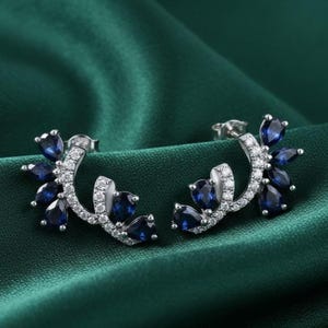 May include: A pair of silver stud earrings with a floral design. Each earring has a curved stem with small, clear stones and clusters of pear-shaped, deep blue gemstones. The earrings are displayed on a dark green satin surface.
