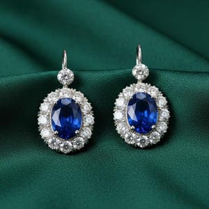 May include: A pair of elegant earrings featuring a large, oval, deep blue gemstone surrounded by a halo of smaller, clear gemstones. The earrings have a silver-colored hook and are set against a dark green background.