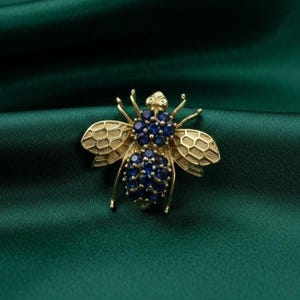 Puede incluir: Broche de abeja dorada con detalles intrincados. Las alas tienen un patrón de panal y el cuerpo está adornado con numerosas piedras preciosas azules. La broche se encuentra sobre un fondo verde oscuro y sedoso.