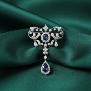 May include: An ornate silver brooch with a central oval sapphire and surrounding clear gemstones. The brooch features a teardrop sapphire pendant and is set against a dark green fabric.