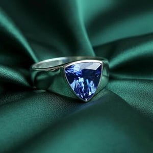 May include: A silver ring featuring a triangular, deep blue gemstone. The ring is set against a backdrop of dark green fabric, creating a contrast that highlights the ring's design. The gemstone has a faceted surface.