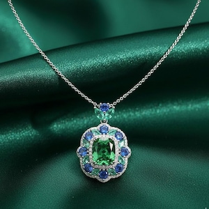Puede incluir: Un collar de plata con un colgante que presenta una gran piedra preciosa verde esmeralda cuadrada, rodeada de piedras azules y verdes más pequeñas. El colgante está engastado en un diseño floral y la cadena es delicada.