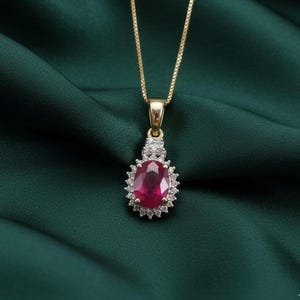 Puede incluir: Un collar de oro con un colgante con un gran rubí ovalado rodeado de pequeños diamantes. El rubí es de color rojo intenso y los diamantes brillan. El collar se exhibe sobre una tela verde oscuro.