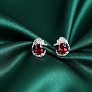 May include: A pair of silver earrings with oval, deep red gemstones. Each earring features a halo of small, clear stones and a decorative top. The earrings are displayed on a dark green, silky fabric.