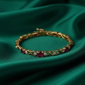 May include: A gold bracelet with alternating oval-cut red gemstones and small round clear gemstones. The bracelet is set against a dark green, silky background.
