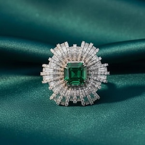 Op de afbeelding: Een opvallende ring met een grote, vierkant geslepen smaragd als middensteen. De smaragd wordt omringd door een halo van kleinere, baguette-geslepen diamanten, wat een zonnestraaleffect creëert. De ring is gezet op een zilveren band.
