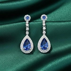 May include: A pair of drop earrings with a pear-shaped, deep blue gemstone at the base, encircled by smaller, clear stones. The earrings have a post and dangle design with small, round, clear stones connecting the top and bottom sections.