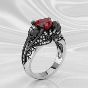 May include: A gothic-style ring featuring a heart-shaped red gemstone. The ring has a black skull design on each side of the gemstone, with intricate detailing and small, clear accent stones. The band is silver-colored.