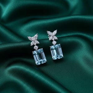 May include: A pair of elegant earrings with a butterfly design. Each earring features a rectangular, light blue gemstone suspended below a cluster of small, clear stones. The earrings are set against a dark green, satin-like fabric.