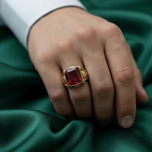 May include: A gold ring featuring a large, rectangular, red gemstone. The ring is worn on a hand, with a white shirt sleeve in the background. The ring's setting has a decorative design.